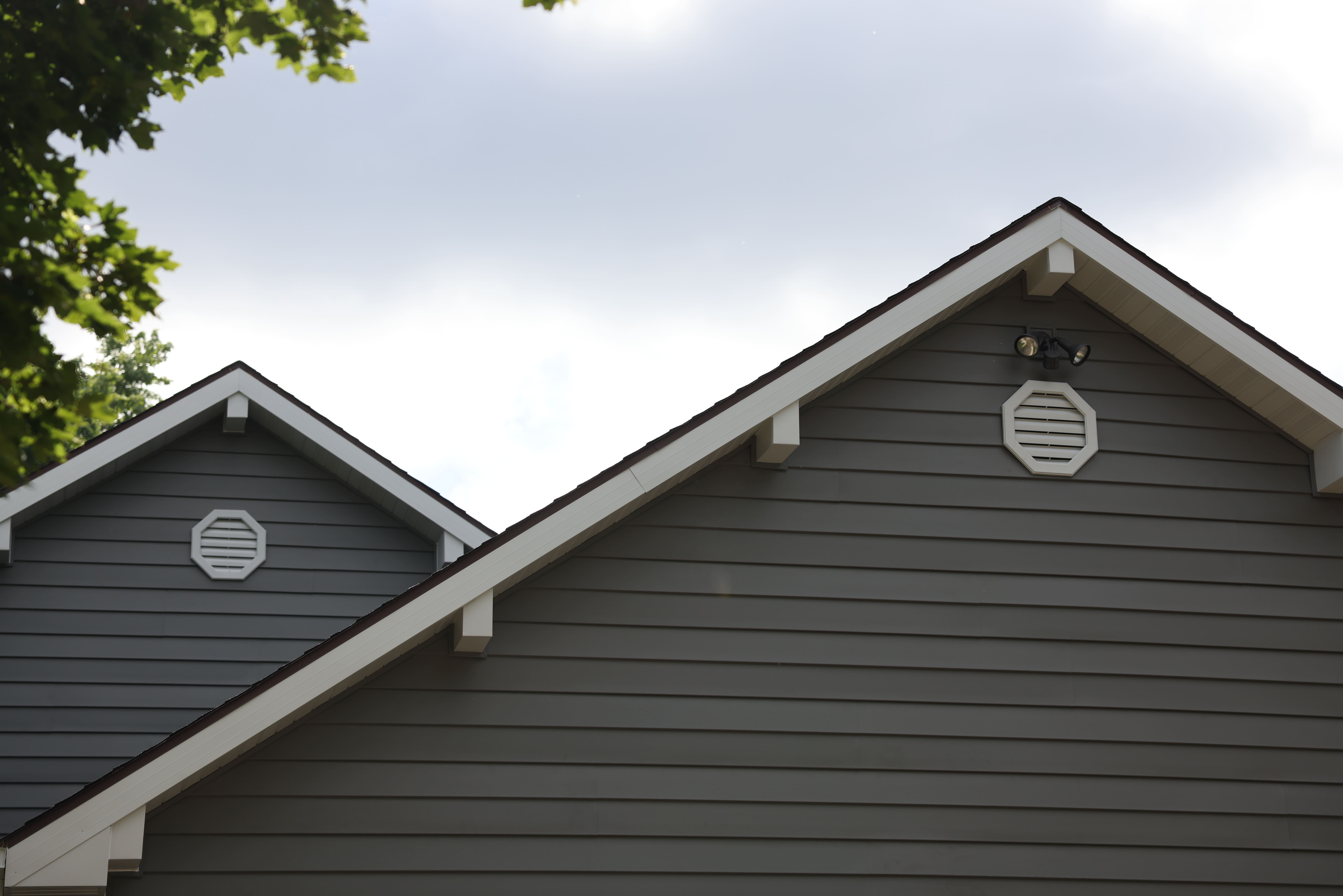 Two gray house gables with white trim and octagonal vents under a cloudy sky with tree leaves framing the top corners.