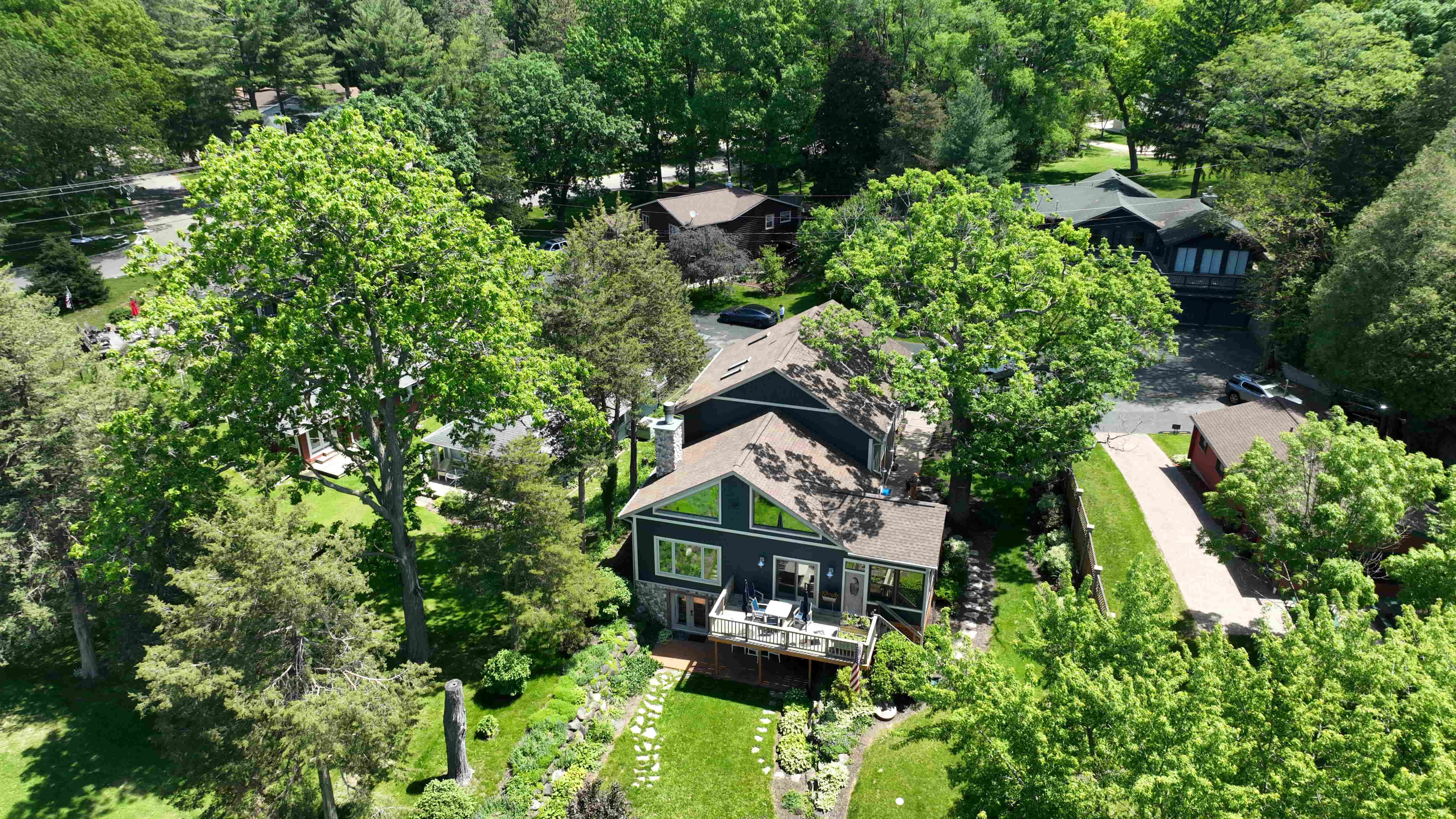 Aerial view of a dark gray house with a large deck, surrounded by lush green trees and a well-maintained yard.